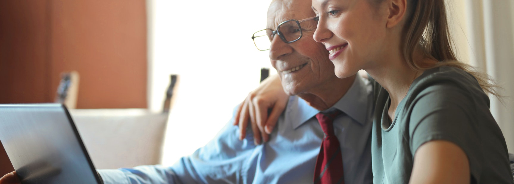 senior citizen man and young woman smiling with one arm around his shoulder while they look at a laptop